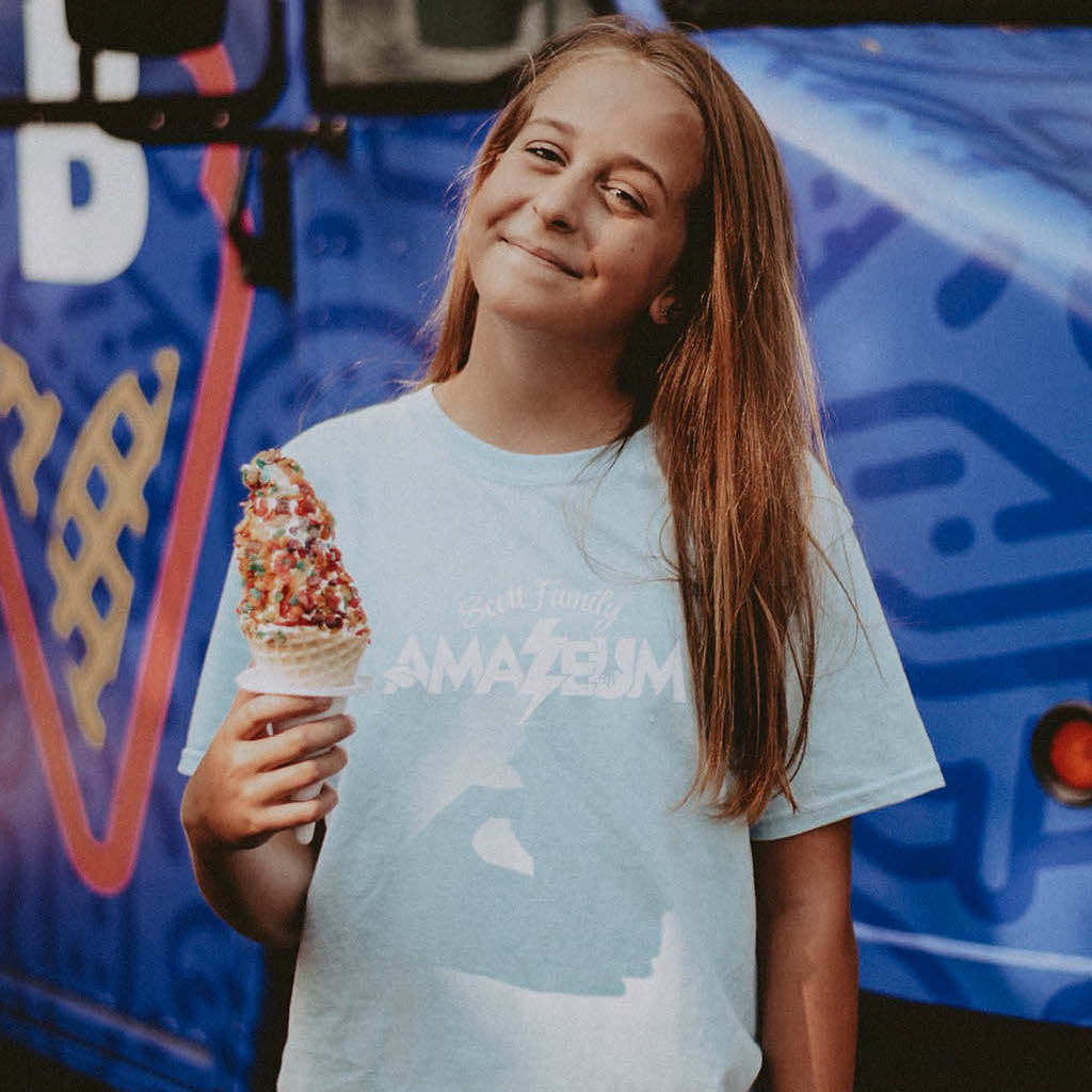 Girl stands with ice cream cone wearing a blue shirt that reads, "Scott Family Amazeum" on it.
