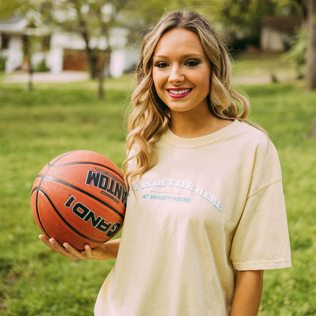 Girl holds basketball and wears a light yellow shirt that reads, "Life is Better Here at Varsity House."