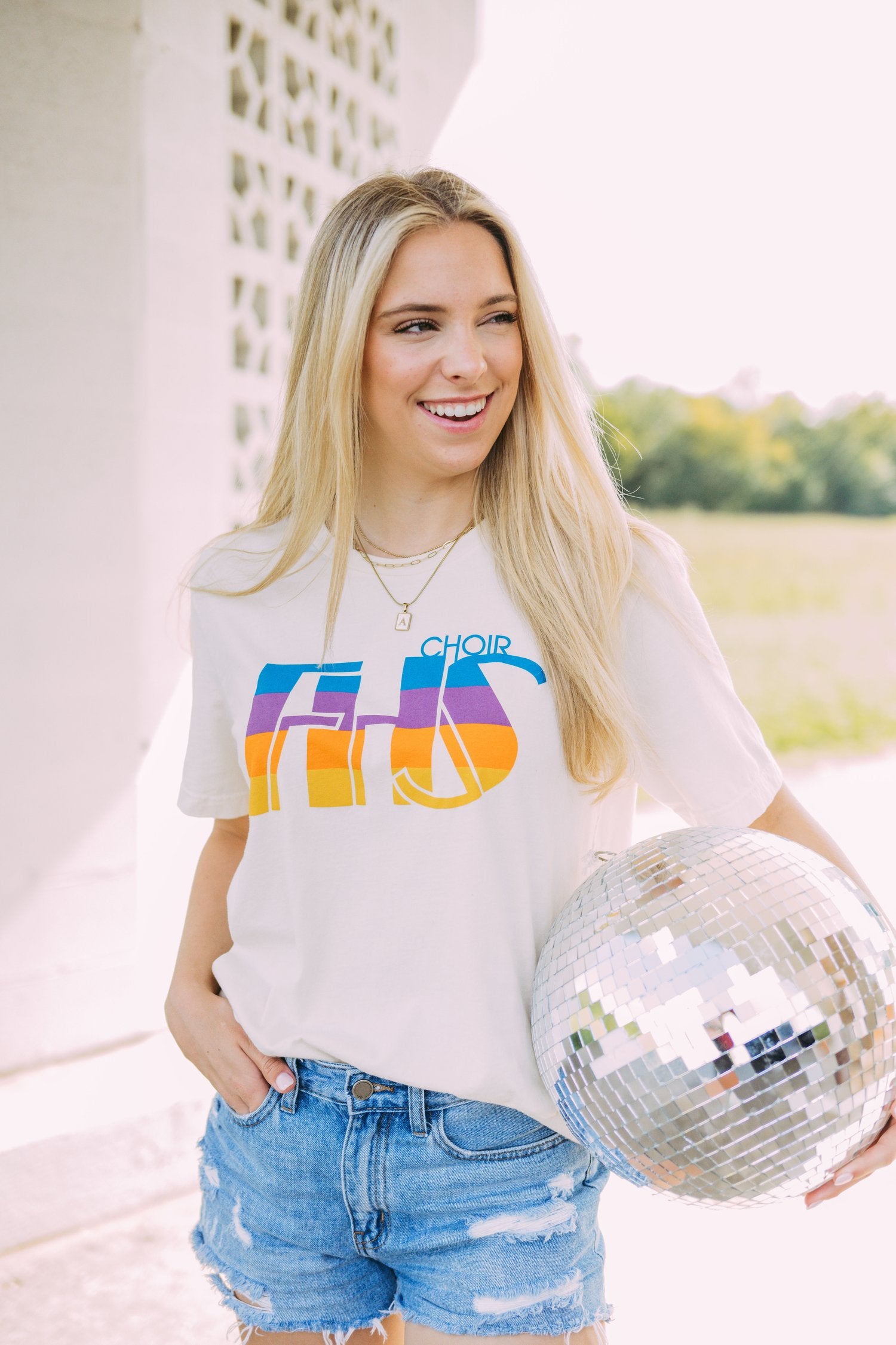 Girl stands in field and holds a disco ball. She wears a white tshirt with "FHS Choir" written across it.