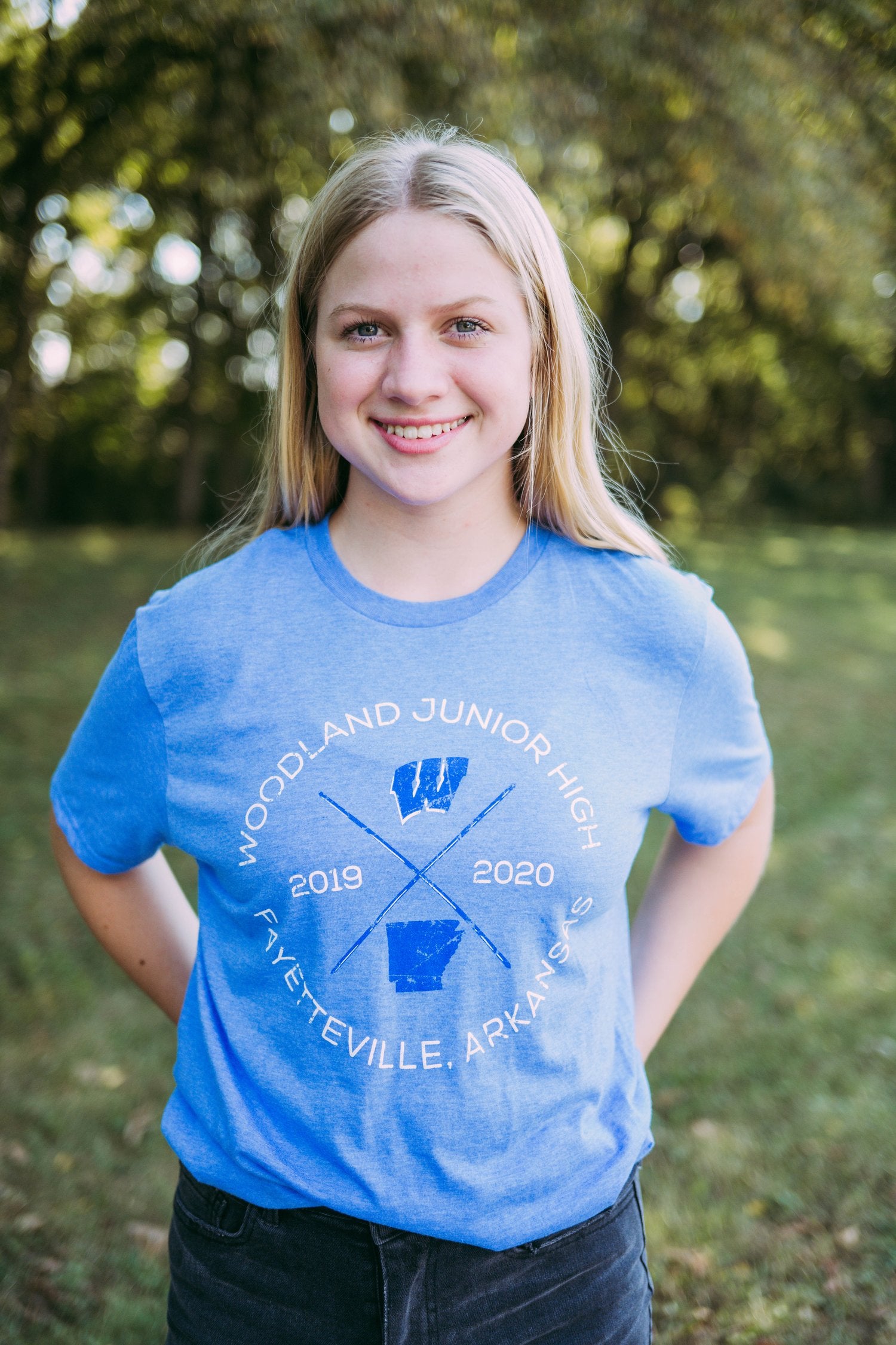 Girl stands in field wearing a blue tshirt that reads, "Woodland Junior High Fayetteville, Arkansas." Shirt has an outline of the state of Arkansas and school logo on it.