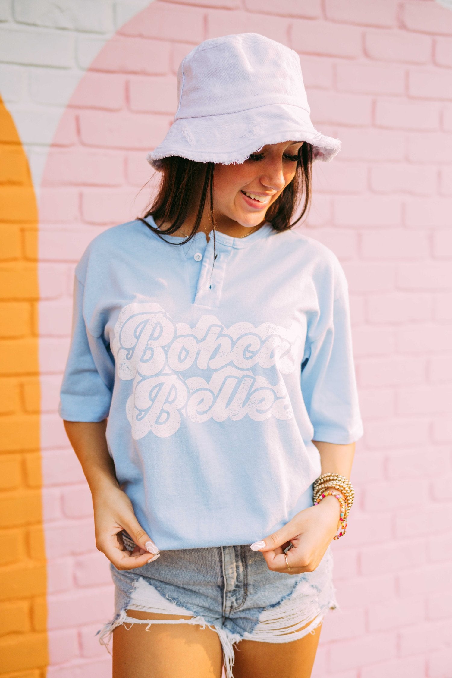 Girl stands in front of a pink wall wearing a light blue henley tshirt that reads, "Bobcat Belles."
